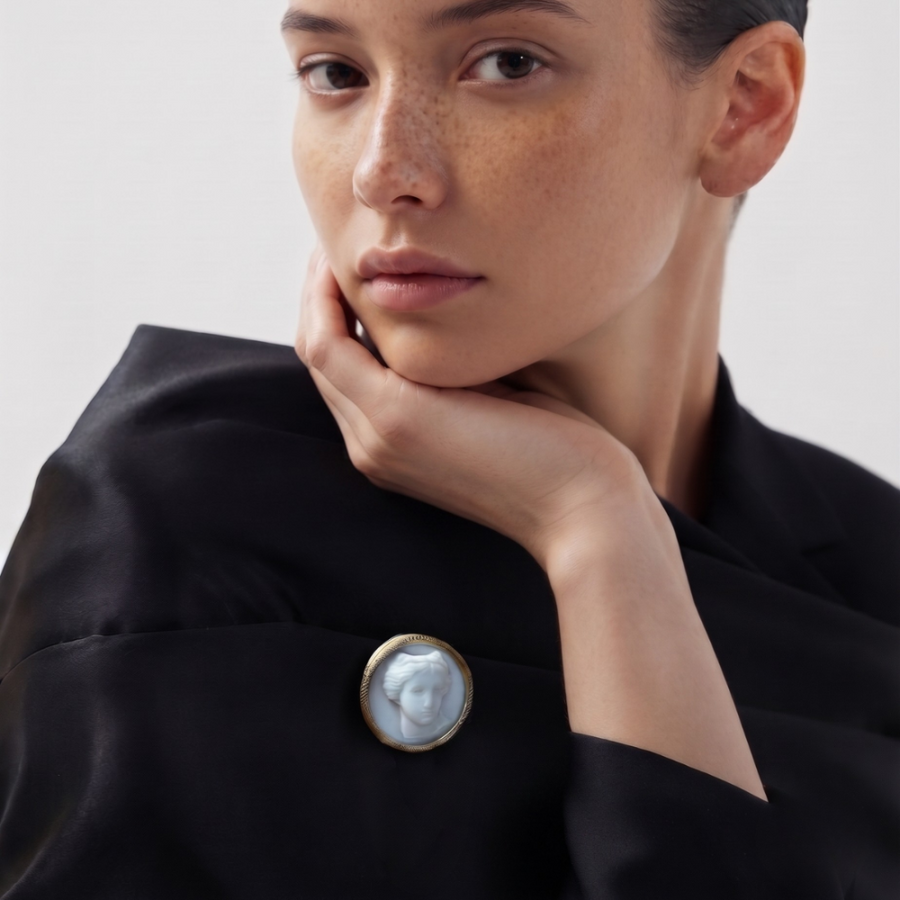 A close-up of a model wearing a black satin jacket with a prominent round button featuring a carved white shell in a gold-tone frame.