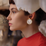 A close-up side profile of a woman wearing a round cameo-style earring with a white face motif and gold-toned border, paired with a red top and white head covering.