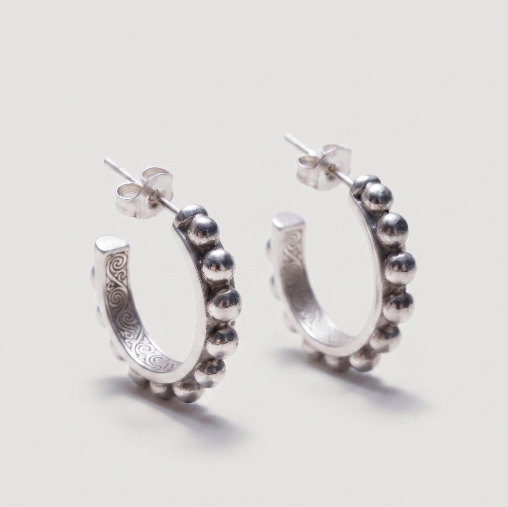 A pair of sterling silver huggie hoop earrings with a beaded outer border and engraved details on the inner band, shown on a light gray background.
