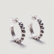 A pair of sterling silver huggie hoop earrings with a beaded outer border and engraved details on the inner band, shown on a light gray background.