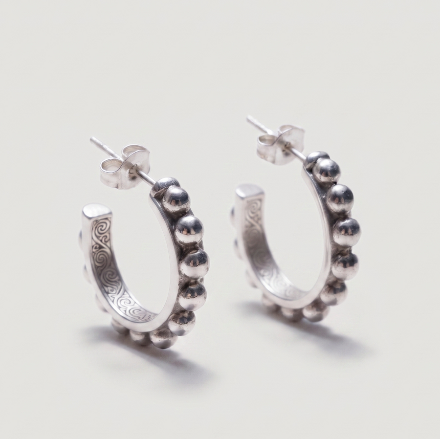 A pair of sterling silver huggie hoop earrings with a beaded outer border and engraved details on the inner band, shown on a light gray background.