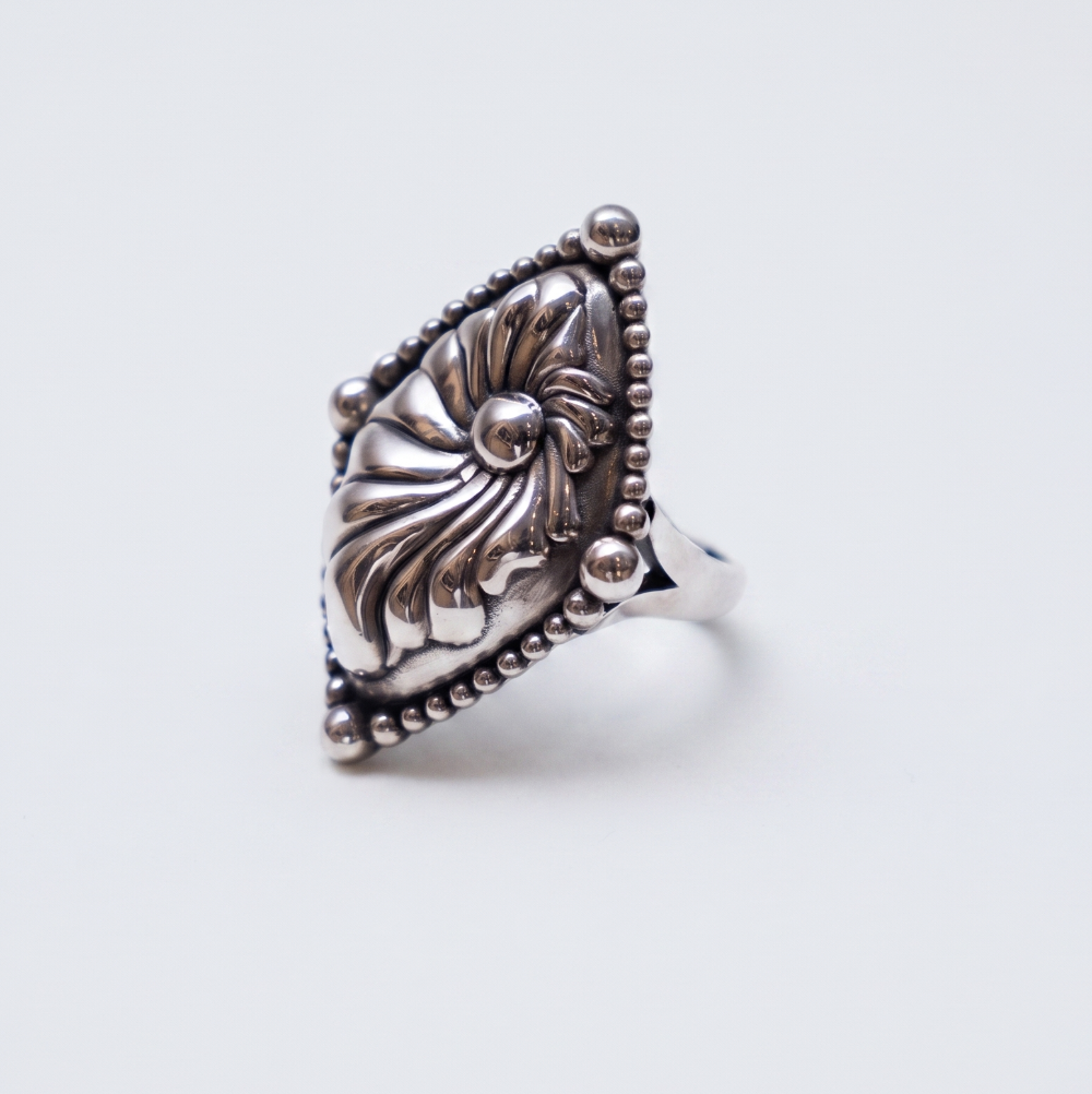 A silver ring with an ornate, carved floral or sunburst design on a shield-like face, bordered by small beads, shown on a light gray background.