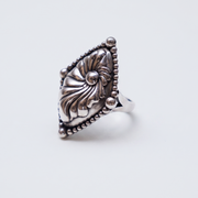 A silver ring with an ornate, carved floral or sunburst design on a shield-like face, bordered by small beads, shown on a light gray background.