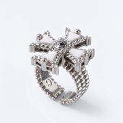 A silver-toned statement ring featuring a decorative cross-shaped top with white inlay sections, a central clear gemstone, and a beaded segmented band, shown on a clean white background.