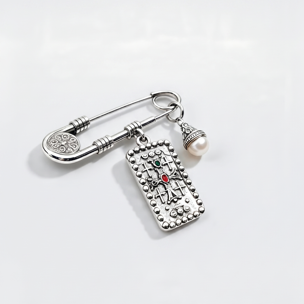 A silver-toned safety pin brooch with a dangling rectangular charm featuring a cross design and a small red gemstone, alongside a dangling pearl bead, on a clean white background.