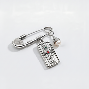 A silver-toned safety pin brooch with a dangling rectangular charm featuring a cross design and a small red gemstone, alongside a dangling pearl bead, on a clean white background.