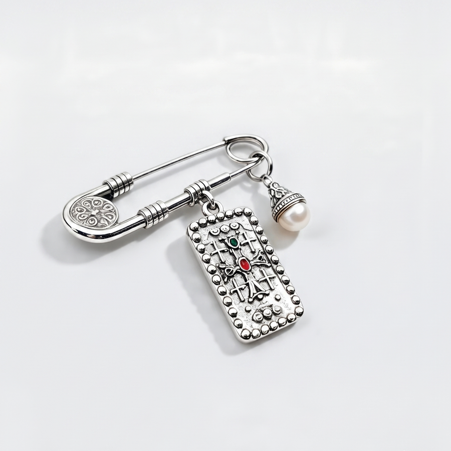 A silver-toned safety pin brooch with a dangling rectangular charm featuring a cross design and a small red gemstone, alongside a dangling pearl bead, on a clean white background.