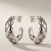 A pair of silver huggie hoop earrings with a textured, woven basketweave pattern on the outer surface and a small flower-shaped stud earring behind each hoop, displayed on a light beige background.
