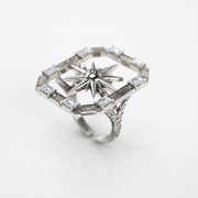 An Art Deco style silver ring featuring a central starburst motif surrounded by a geometric border set with baguette and round clear stones, photographed on a clean white background.