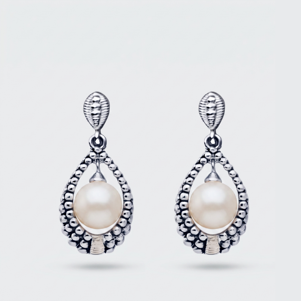 A pair of silver teardrop dangle earrings featuring a central white pearl surrounded by a textured beaded silver border, set against a light gray background.