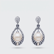 A pair of silver teardrop dangle earrings featuring a central white pearl surrounded by a textured beaded silver border, set against a light gray background.