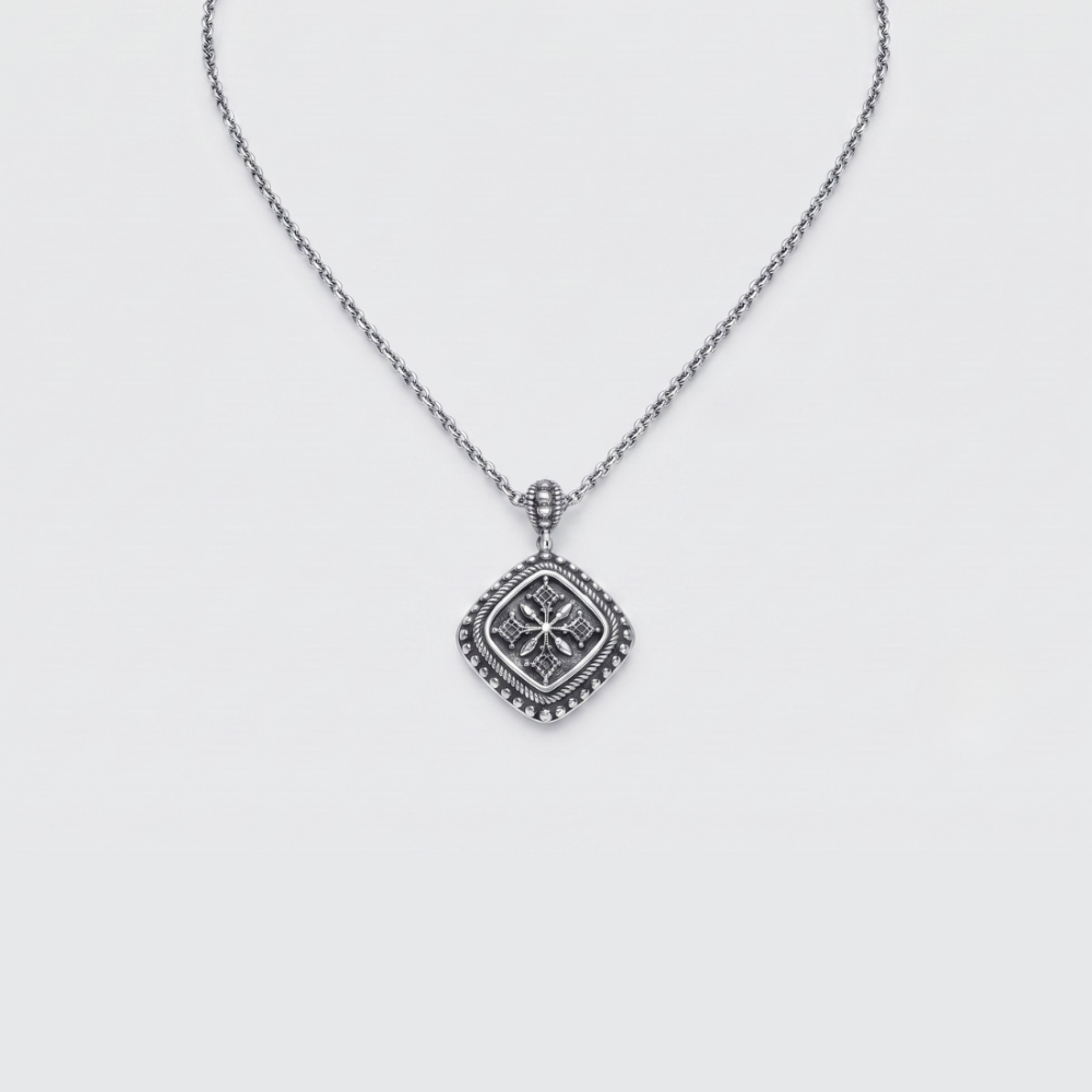 A silver necklace with a square pendant featuring a raised floral or starburst motif and a beaded border, shown on a light gray background.