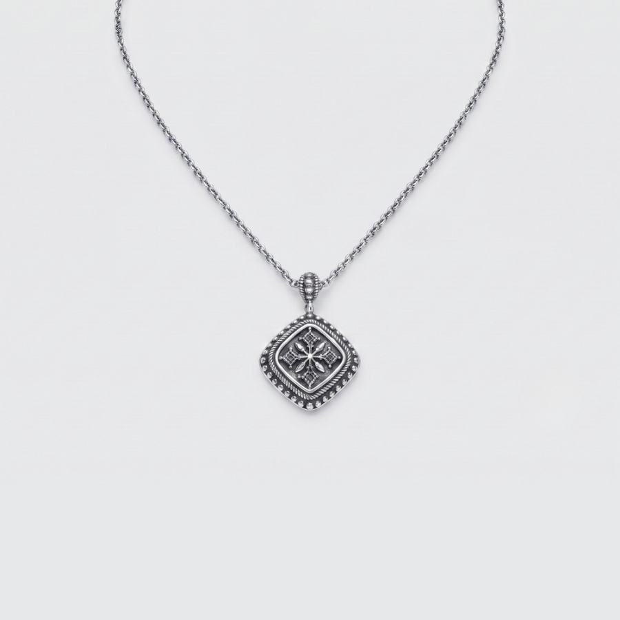A silver necklace with a square pendant featuring a raised floral or starburst motif and a beaded border, shown on a light gray background.