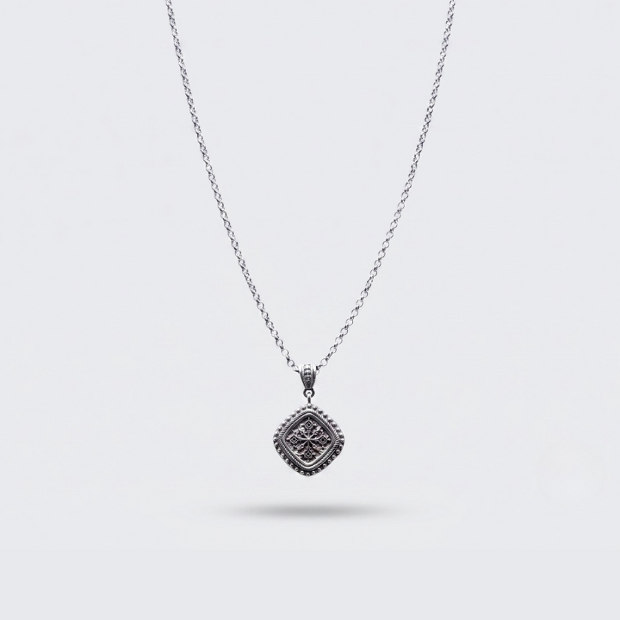 A silver-toned chain necklace with a square pendant featuring an intricate floral or starburst pattern, shown against a light gray background.