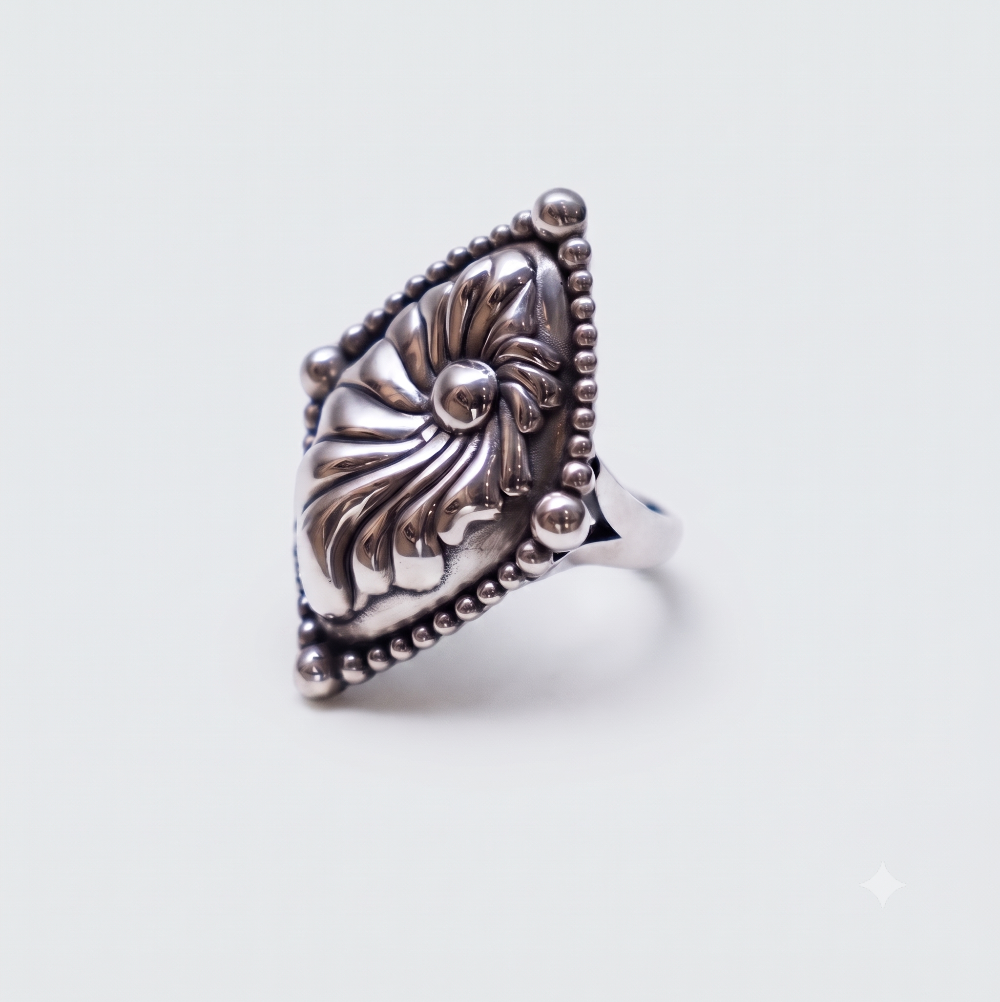 A silver ring featuring an ornate, textured floral or nautilus shell-like centerpiece with a beaded triangular border, shown on a plain light gray background.