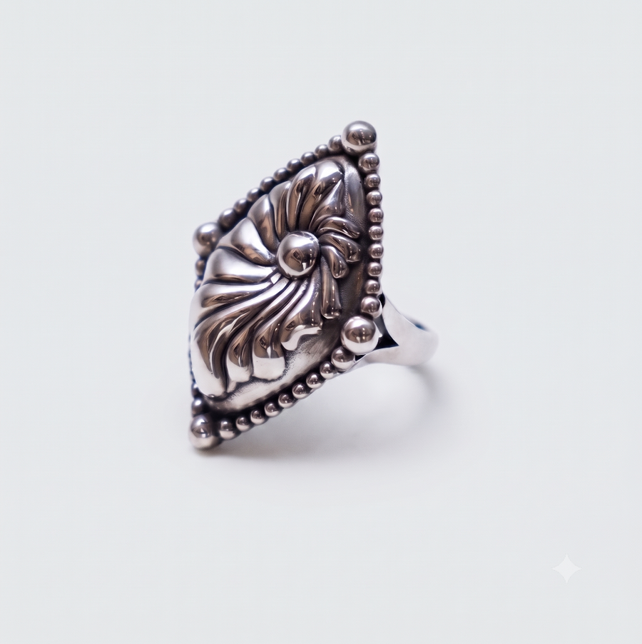 A silver ring featuring an ornate, textured floral or nautilus shell-like centerpiece with a beaded triangular border, shown on a plain light gray background.