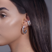 A close-up side profile of a woman with long dark hair wearing a silver crescent-shaped hoop earring adorned with clear rhinestones, along with a pearl necklace.