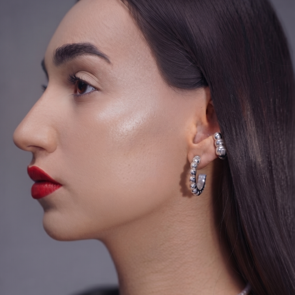 A close-up side profile of a woman with dark hair wearing a pearl necklace and matching drop earrings with sparkling clear stones, against a soft gray background.