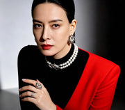 A woman wearing a striking red and black color-blocked dress, styled with a double-strand pearl necklace and statement rings, posing in a dramatic studio setting.