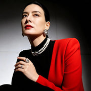 A woman seated in a dramatic studio portrait wearing a bright red blazer over a black top, with layered pearl necklaces, statement earrings, and rings. The background is dark with a spotlight effect.
