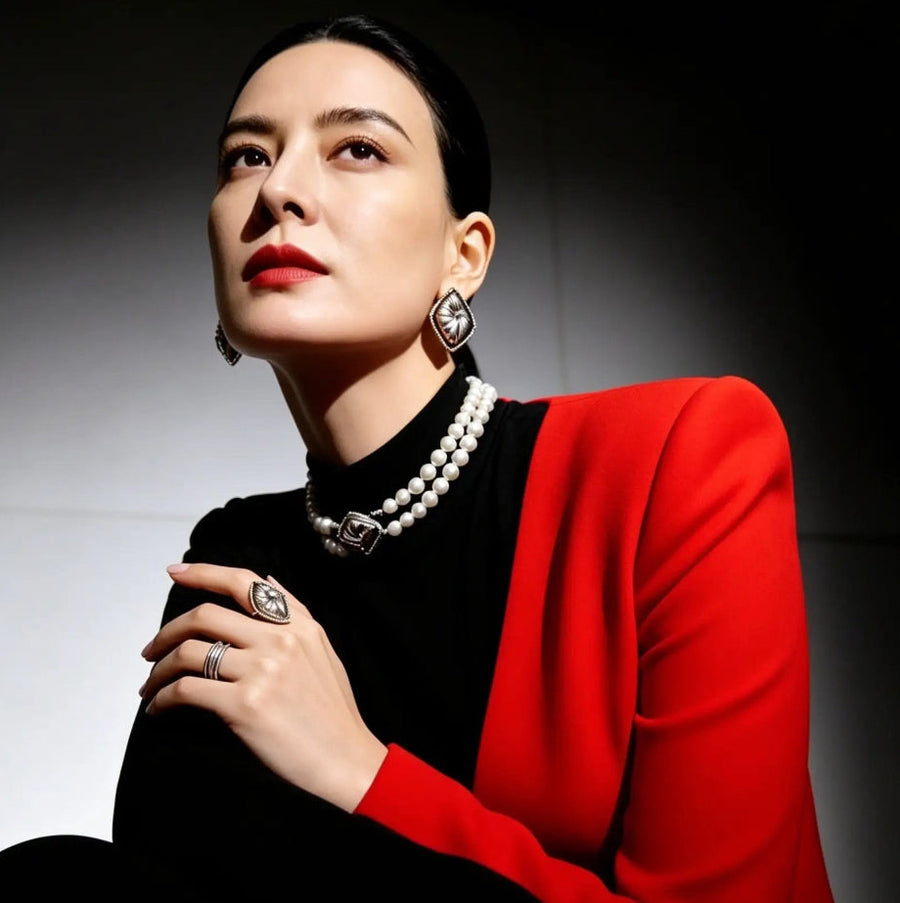 A woman seated in a dramatic studio portrait wearing a bright red blazer over a black top, with layered pearl necklaces, statement earrings, and rings. The background is dark with a spotlight effect.