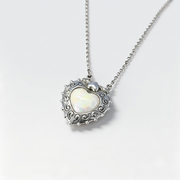 A silver heart-shaped pendant necklace with an iridescent white opal center and ornate scrollwork, shown on a delicate silver chain against a light gray background.