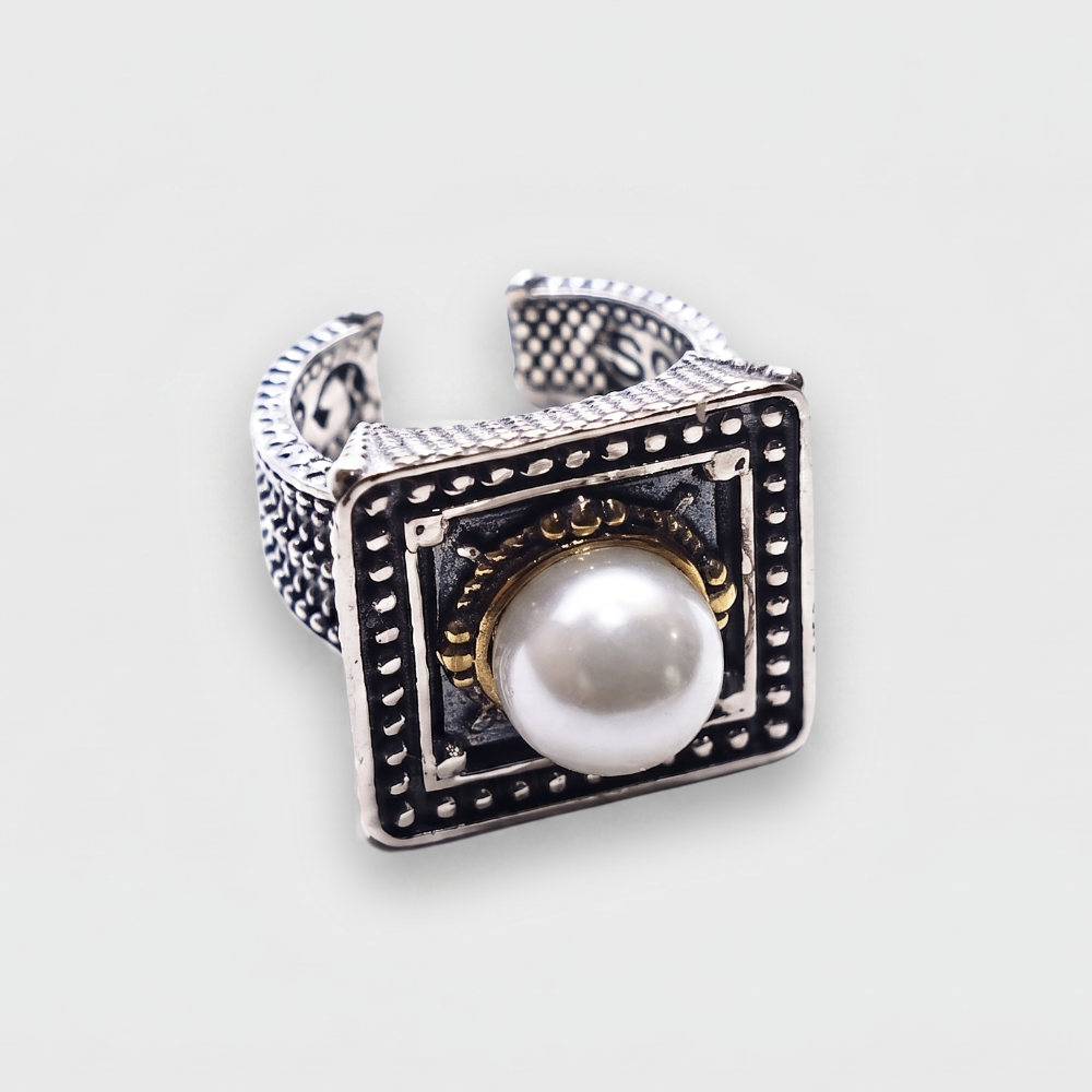 A silver-toned square ring with a central white pearl, black oxidized detailing, and small gold accents, shown on a light gray background.