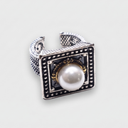 A silver-toned square ring with a central white pearl, black oxidized detailing, and small gold accents, shown on a light gray background.