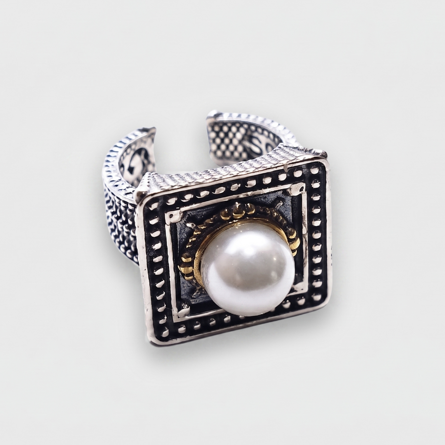 A silver-toned square ring with a central white pearl, black oxidized detailing, and small gold accents, shown on a light gray background.