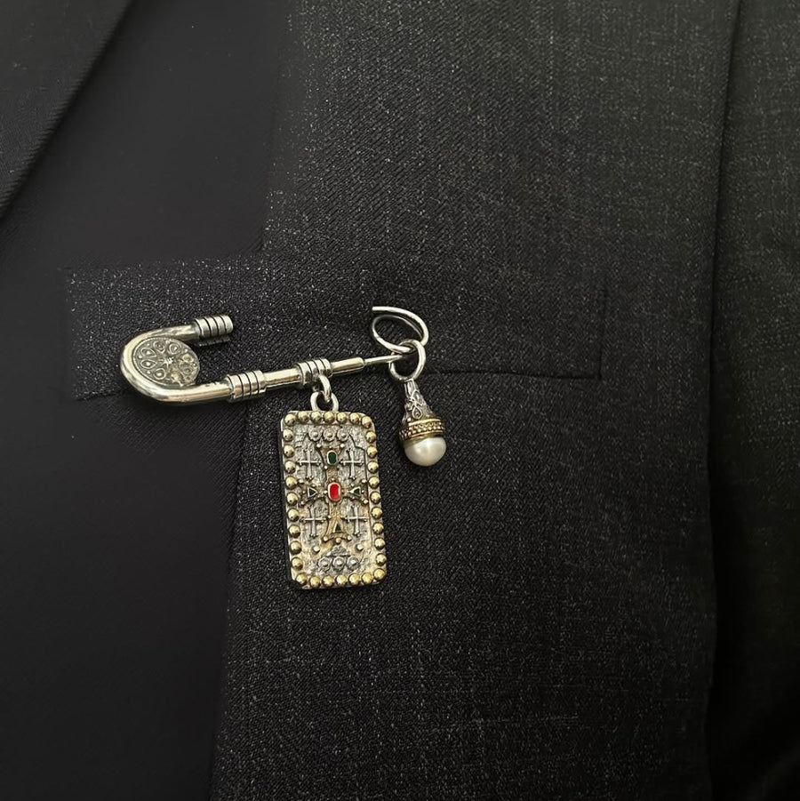 A silver-toned men's lapel pin brooch featuring a rectangular enamel-embellished plate with a small red center stone, a key-shaped pin, and a dangling pearl bead, pinned to a black suit jacket.