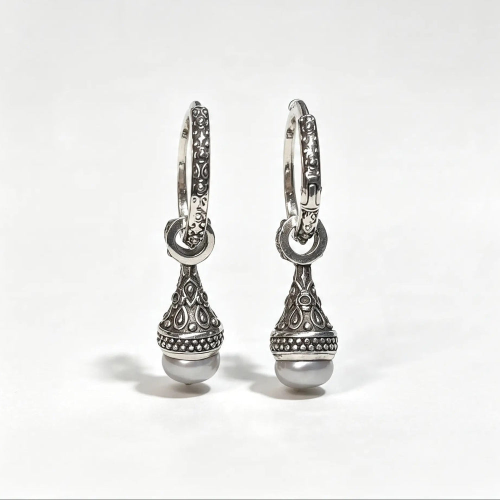 A pair of silver huggie hoop earrings with ornate filigree detailing and a dangling teardrop-shaped charm ending in a round pearl, shown on a light gray background.