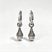 A pair of silver huggie hoop earrings with ornate filigree detailing and a dangling teardrop-shaped charm ending in a round pearl, shown on a light gray background.