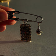 A silver-toned safety pin brooch with a rectangular engraved pendant and a small teardrop charm, held by a hand against a warm-lit gray surface.