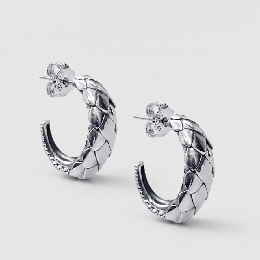 A pair of silver hoop earrings with a textured, woven or scaled pattern and post-back closures, shown on a light gray background.