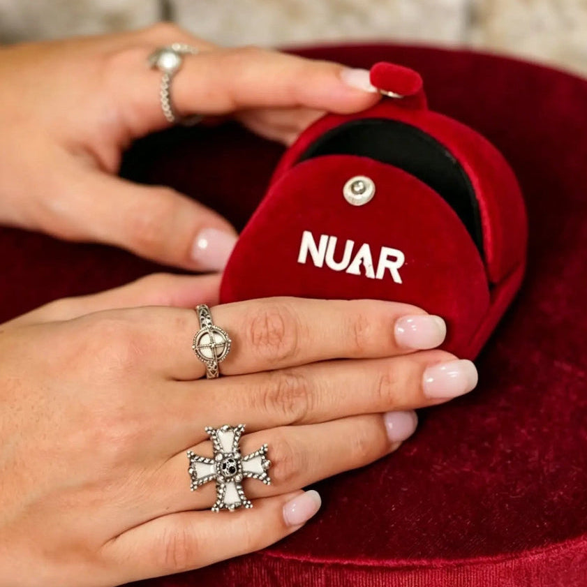 A close-up of hands holding an open red velvet ring box with the brand name NUAR printed in white on the front. The hands have light pink manicured nails and are wearing silver rings, including a prominent cross-shaped ring with clear stones.