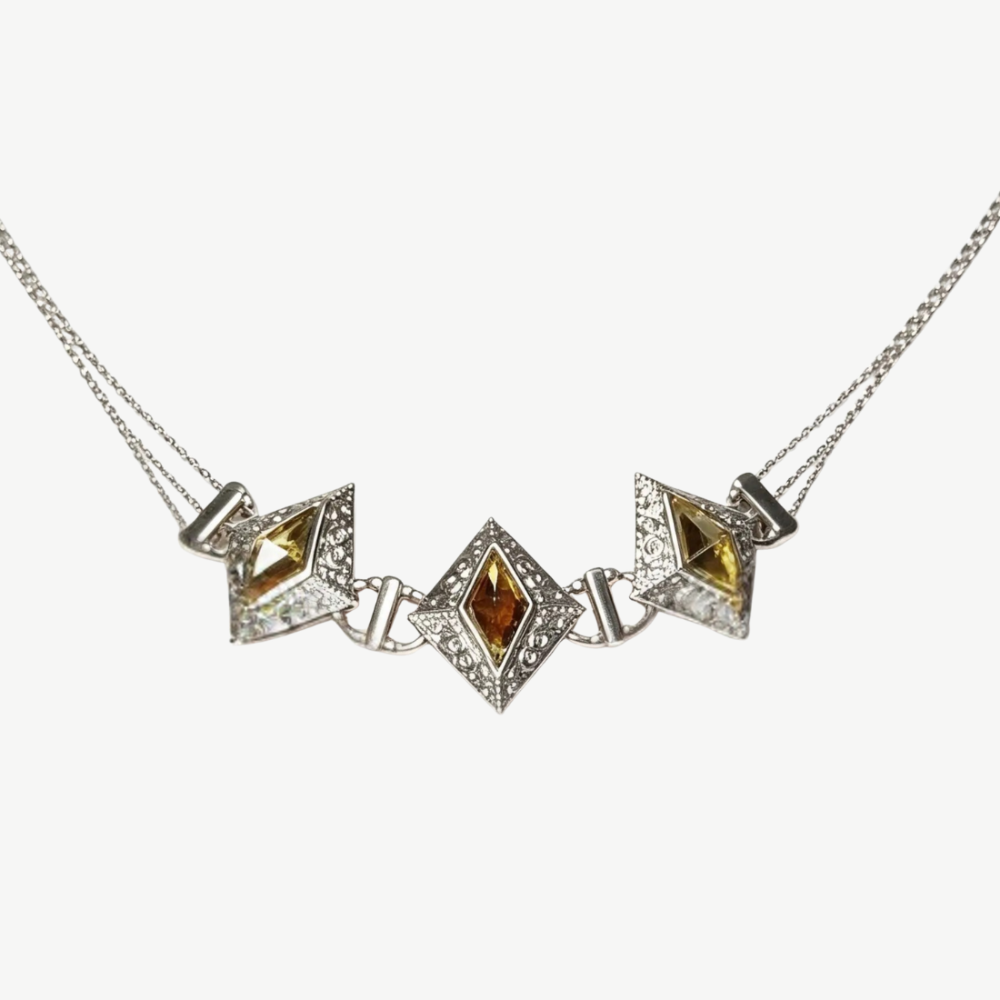 A silver-toned geometric necklace featuring three diamond-shaped puffed centers, with the middle and right centers containing yellow gemstones and the left center containing an amber-brown gemstone, all set with small clear stones on a light gray background.
