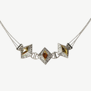 A silver-toned geometric necklace featuring three diamond-shaped puffed centers, with the middle and right centers containing yellow gemstones and the left center containing an amber-brown gemstone, all set with small clear stones on a light gray background.