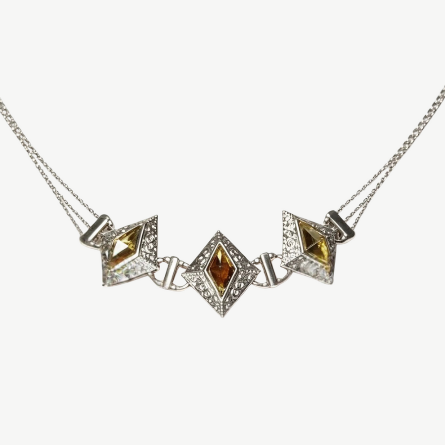 A silver-toned geometric necklace featuring three diamond-shaped puffed centers, with the middle and right centers containing yellow gemstones and the left center containing an amber-brown gemstone, all set with small clear stones on a light gray background.