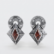 A pair of silver-toned drop earrings with textured circular tops and diamond-shaped pendants featuring a central dark red gemstone, shown on a light gray background.