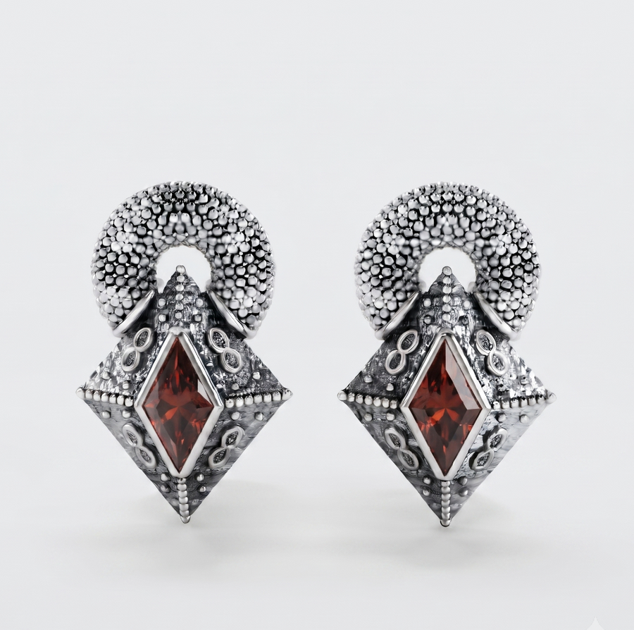 A pair of silver-toned drop earrings with textured circular tops and diamond-shaped pendants featuring a central dark red gemstone, shown on a light gray background.