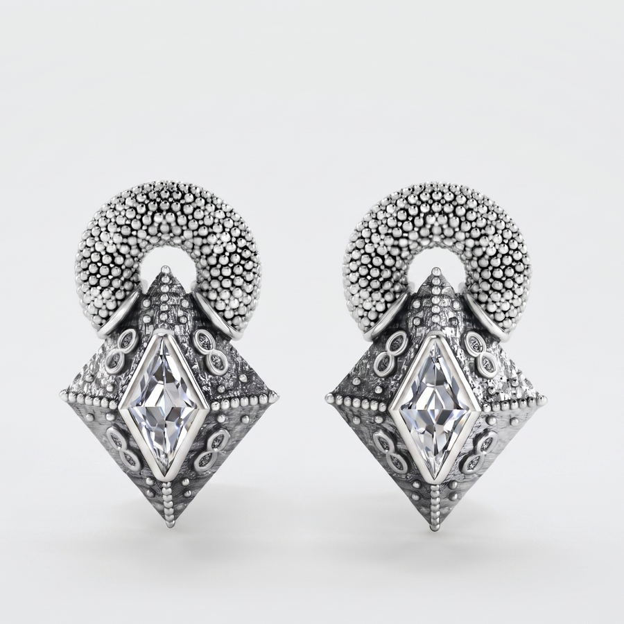 A pair of silver-toned drop earrings with a textured circular top and a faceted diamond-shaped pendant featuring a central clear gemstone, shown on a light gray background.