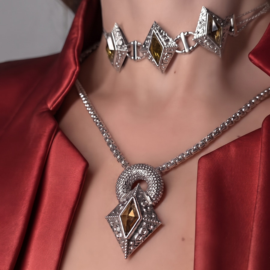 Silver-toned jewelry set on a model, featuring a choker necklace and a matching pendant necklace with diamond-cut amber-colored stones and textured silver settings, worn with a red satin jacket.