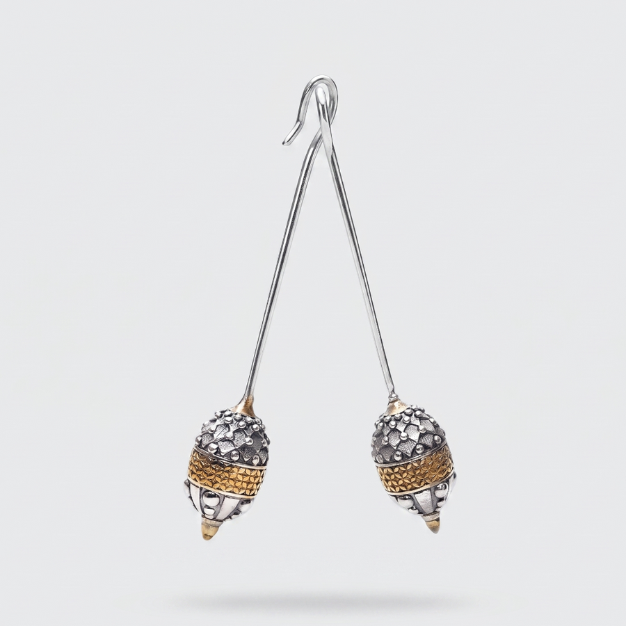 A pair of dangling earrings with long silver-tone wire stems and acorn-shaped drops. The acorns are textured with silver pavé stones and a band of gold-tone detailing, shown against a light gray background.