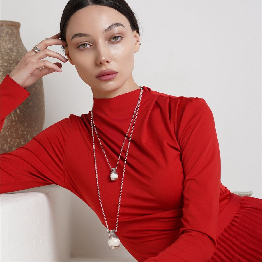 A woman wearing a bright red long-sleeve mock-neck dress and layered silver necklaces with pearl-like pendants, posed indoors against a light neutral background.