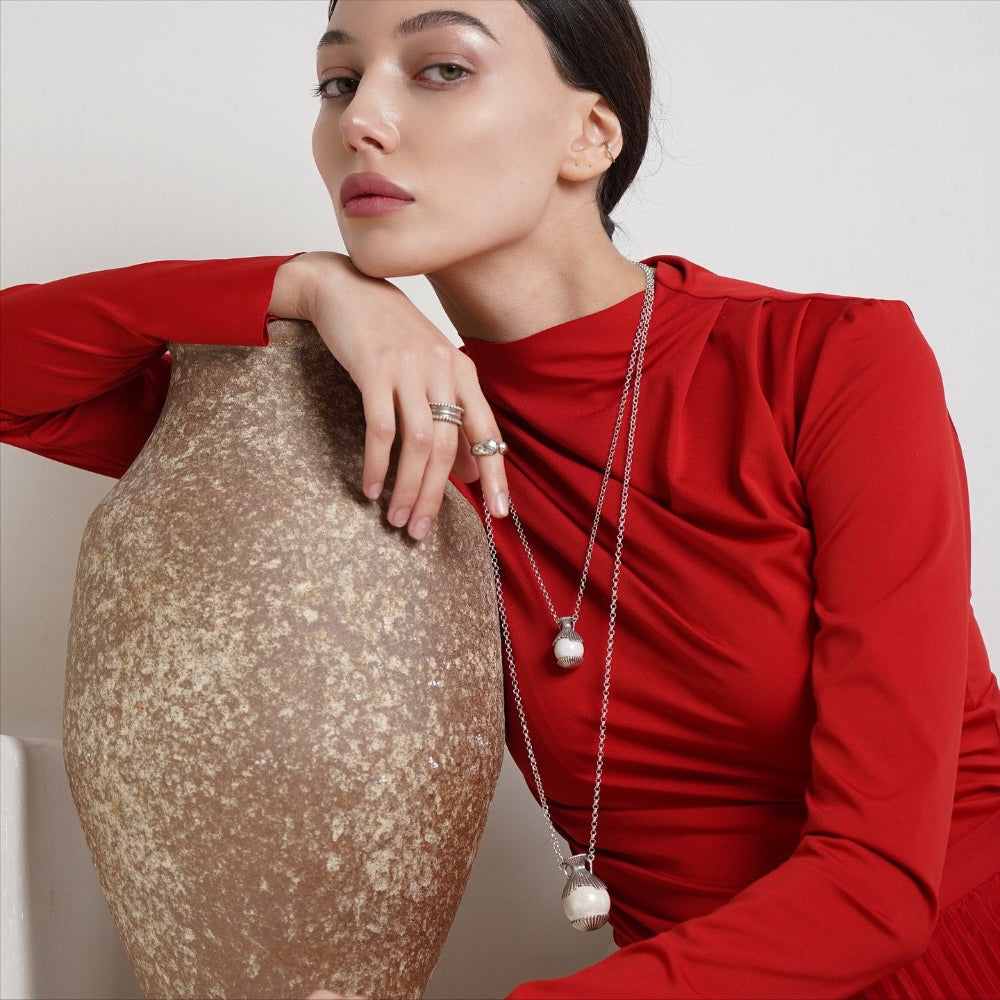 A woman in a red long-sleeve top models a silver double-layered necklace with pearl-like oval pendants, leaning against a textured brown ceramic vase.