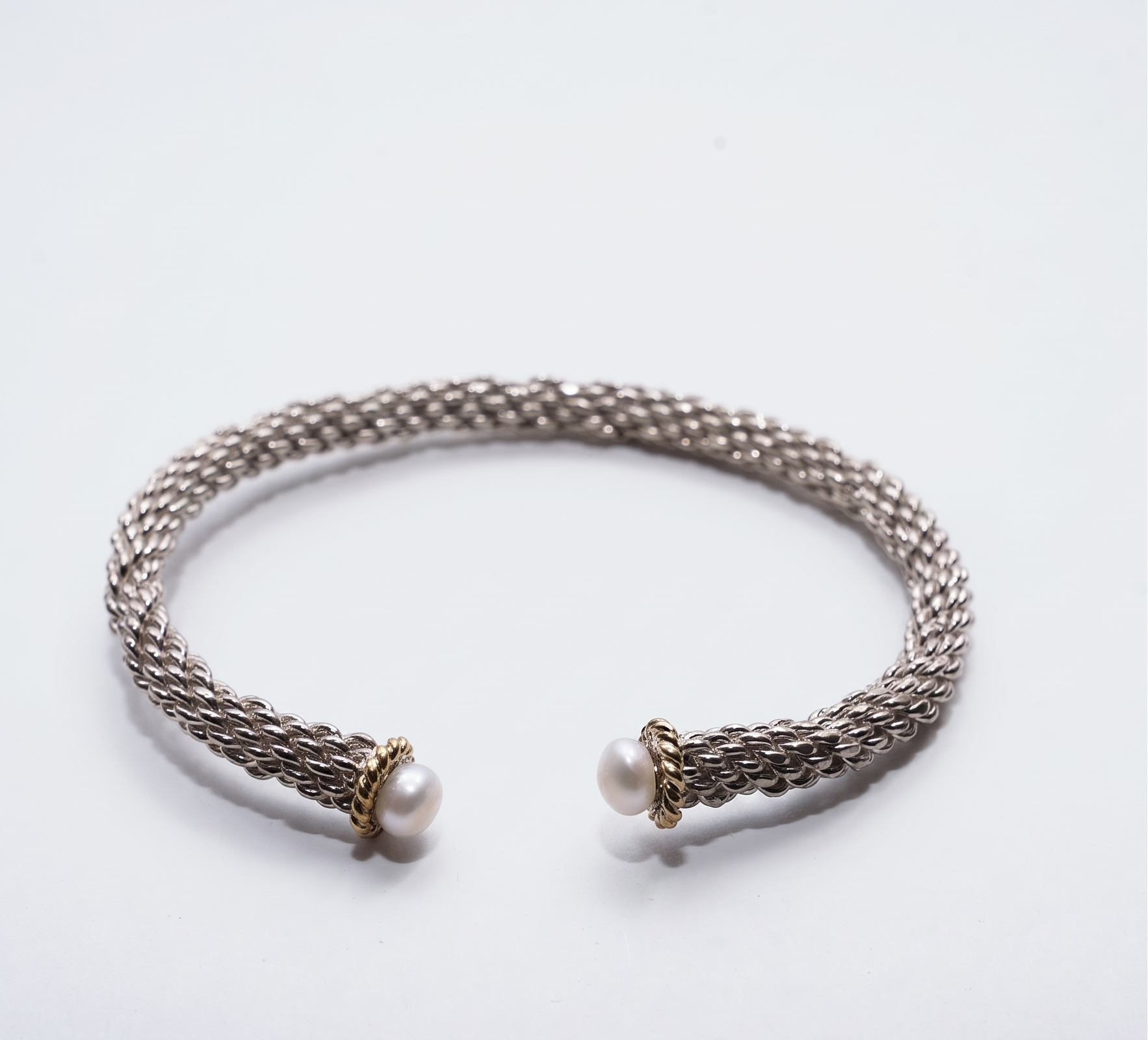 A silver-toned open cuff bracelet with a braided mesh texture and two small white pearl accents at the ends, set against a plain white background.