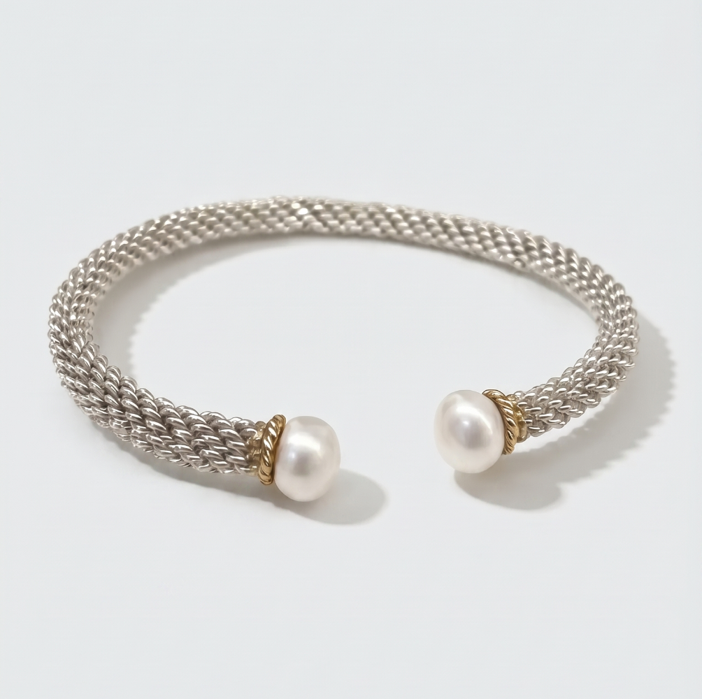 A silver-toned woven cuff bracelet with two white pearl ends and small gold-toned accents, photographed on a clean light gray background.
