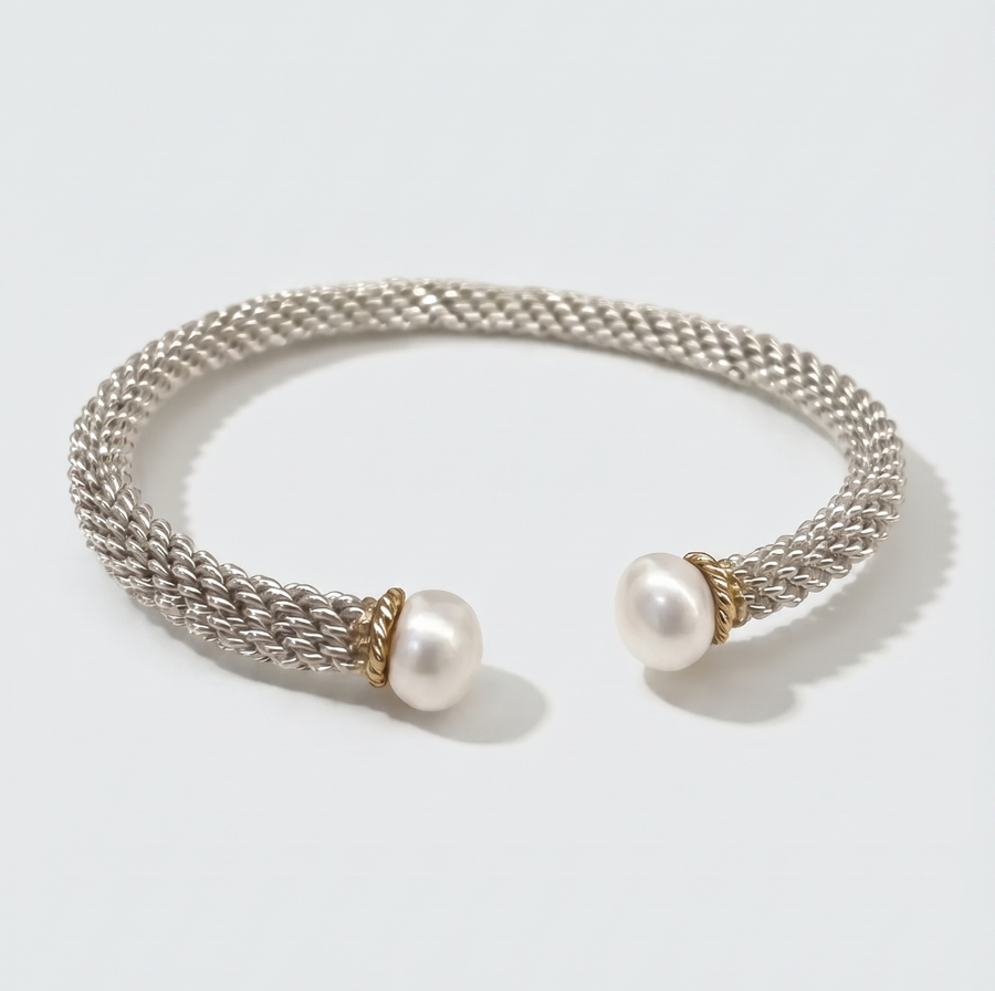 A silver-toned woven cuff bracelet with two white pearl ends and small gold-toned accents, photographed on a clean light gray background.