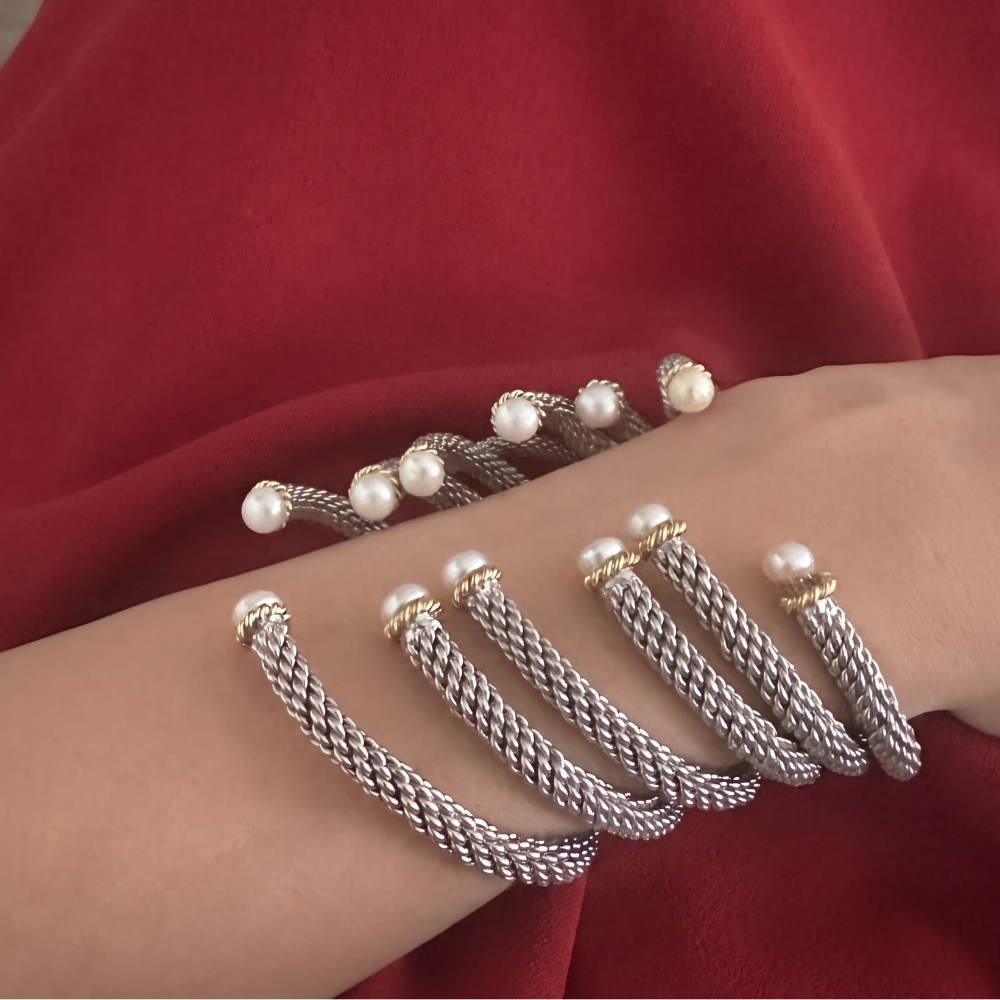 A close-up of a person's wrist wearing two silver cable-wrapped bracelets, one with small white pearls and one with gold and pearl accents, set against a rich red fabric background.