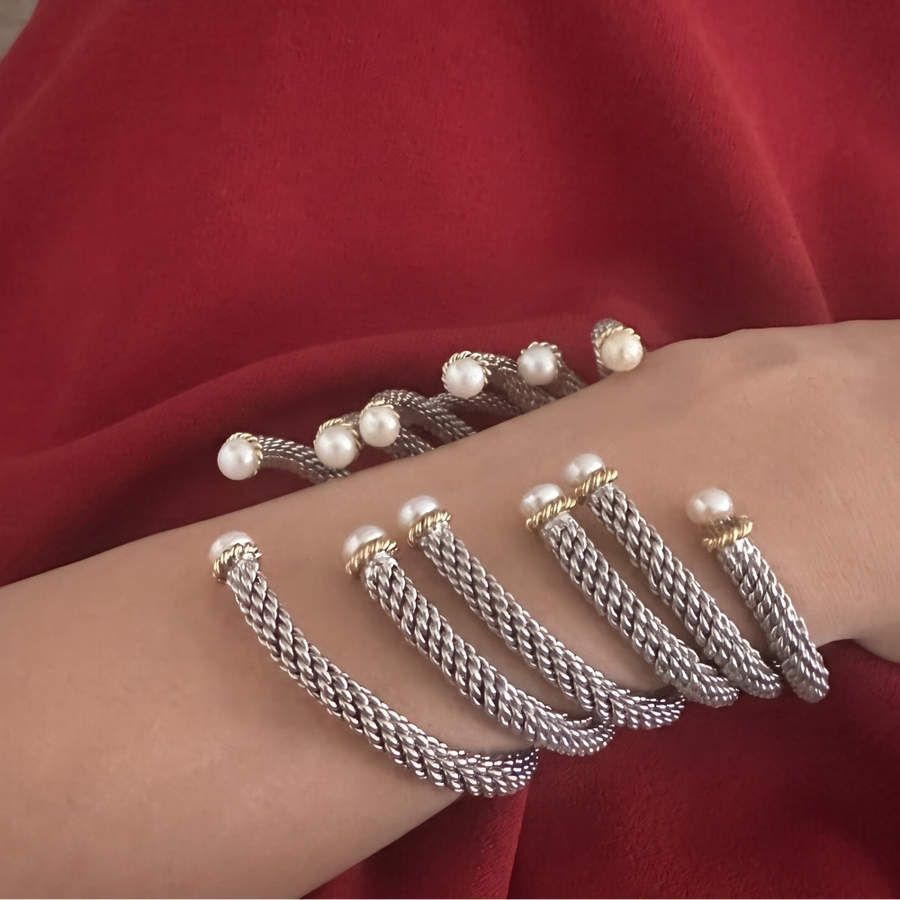 A close-up of a person's wrist wearing two silver cable-wrapped bracelets, one with small white pearls and one with gold and pearl accents, set against a rich red fabric background.